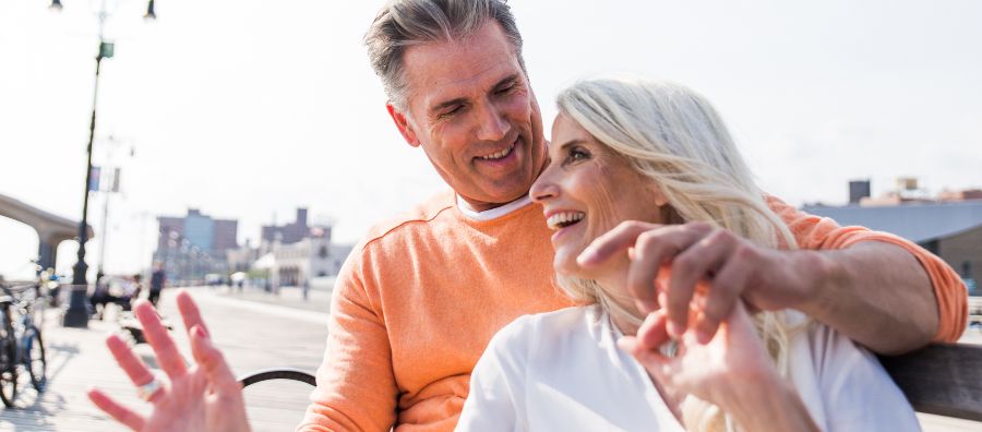 Ein fröhliches Paar beim Dating in Köln über 40 sitzt auf einer Bank am Wasser, lacht und genießt den Moment.